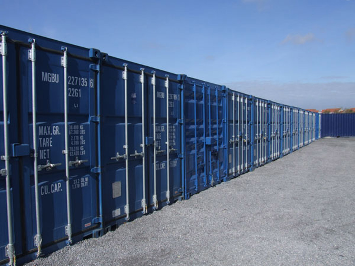 A long row of secure blue 20ft self-storage containers on a gravel yard